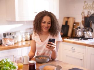 A woman in her kitchen using her phone; smiling
