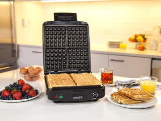 A Krups waffle maker surrounded by breakfast foods on a countertop