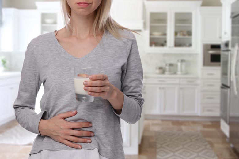 A blonde woman standing in her kitchen with a glass of milk and her hand against her stomach, experiencing discomfort