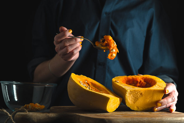 Spoon with seeds in a hand, pumpkin halves on a table on a blue background