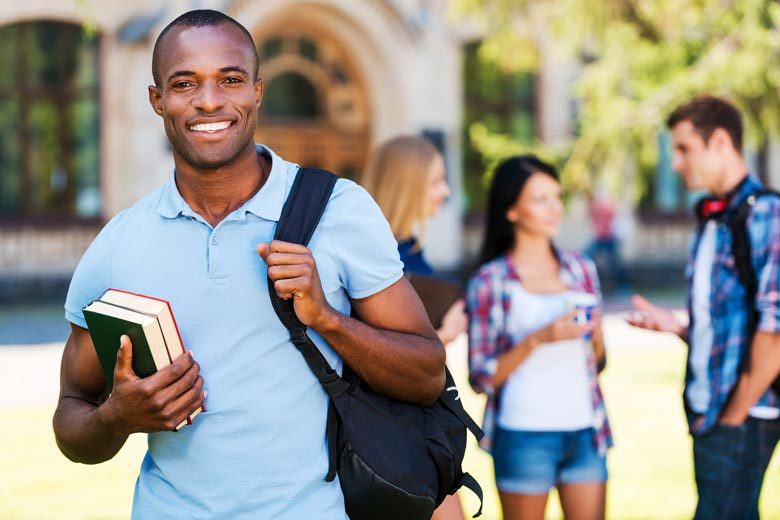 Young African-American male student with books and backpack standing in front of other students, smiling. Shot is taken outdoors with a university building and green trees in the background.