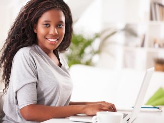 Smiling African-American woman sits at a desk, working on a laptop. She's making eye contact with the camera.