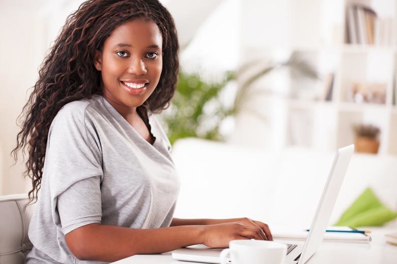 Smiling African-American woman sits at a desk, working on a laptop. She's making eye contact with the camera.