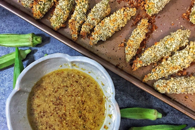 A baking tray of oven baked okra next to a bowl of honey dijon dipping sauce