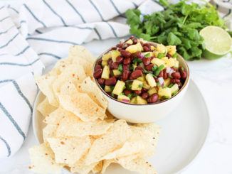 A white bowl filled with pineapple black bean salsa served on a plate with chips. A striped tea towel and bunch of cilantro are in the background.