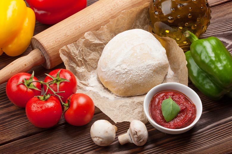 A ball of uncooked pizza dough surrounded by veggies, tomato sauce and a rolling pin