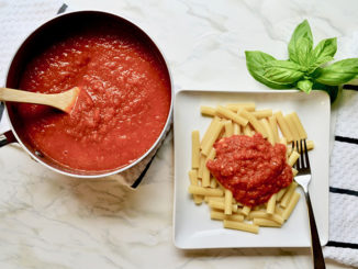 Lagostina Rossella Red Ceramic and Stainless Steel 1.6 Qt. Covered Saucepan with tomato sauce, wooden spoon next to a plate of pasta on a white marble countertop with fresh basil leaves nearby