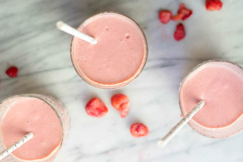 Three pink smoothies with straws on marble countertop sprinkled with raspberries